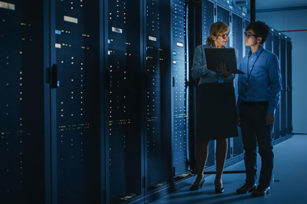 In the Modern Data Center: Engineer and IT Specialist Work with Server Racks, Talking, Doing System Maintenance Check with Laptop Computer.