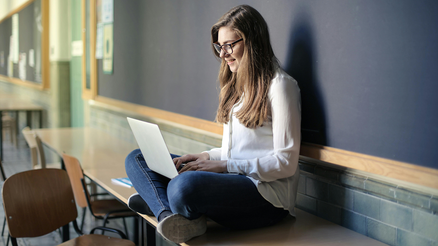 A young women sits on a desk, her laptop on her knees.