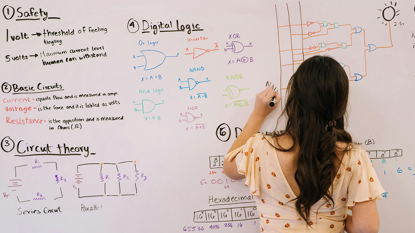 A woman stands at a whiteboard to outline how a computer works, from circuit development to digital logic processing.