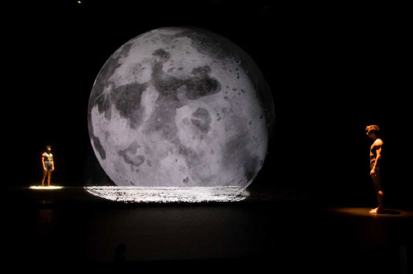Two performing students standing far apart and facing each other, with the giant moon looming in between.