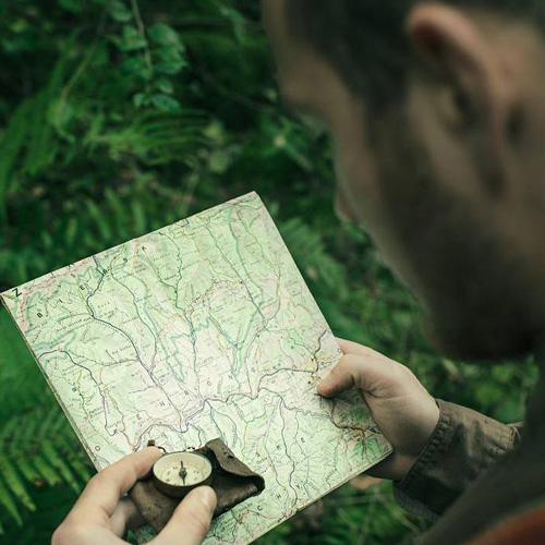 A man stands in the woods, holding a map in one hand and a compass in the other, navigating his surroundings.