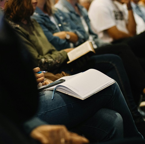 picture of a row of individuals attending a speaker. Individual closest to the camera has a notebook open on their lap with a cell phone and pen.