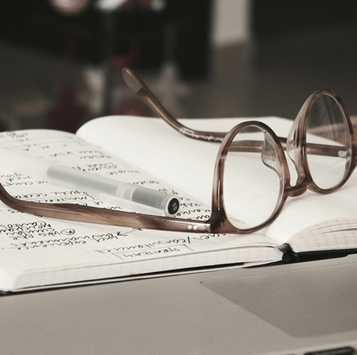 Brown eyeglasses resting upside down on an open notebook with a pen and research notes handwritten on the open page.