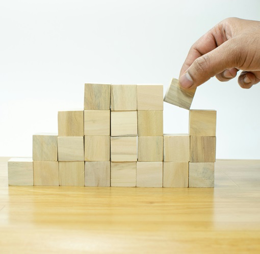 small wooden square blocks being stacked on a table with one hand in the act of adding another block. This signifies providing the building blocks for faculty success