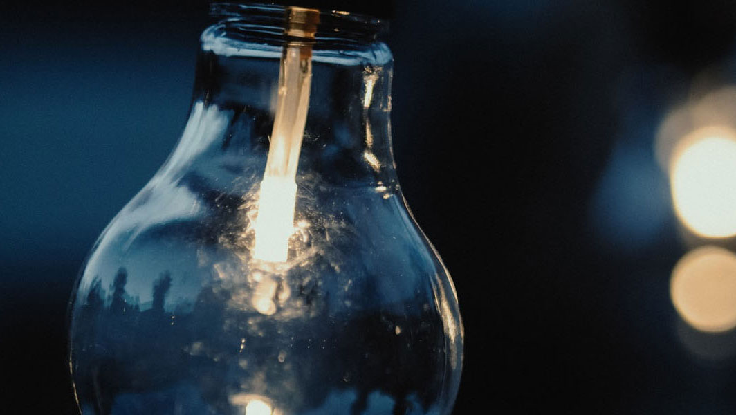 Close-up of a glowing light bulb with a visible filament, set against a dark, blurred background with warm light bokeh in the distance.
