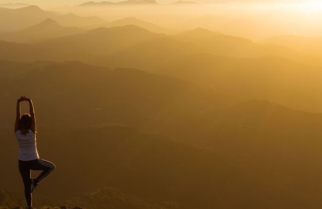 A woman performs yoga on a mountain summit, with breathtaking scenery stretching out in all directions around her.