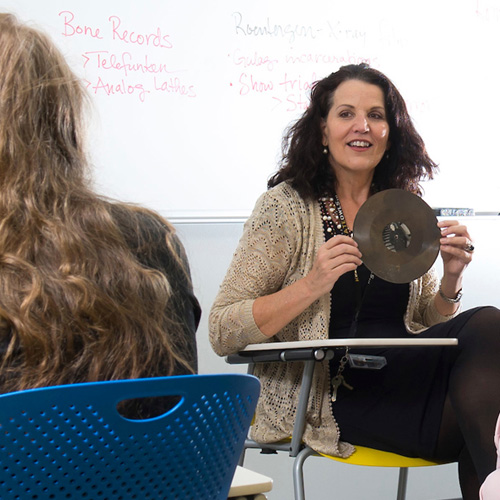 A woman in a chair, holding a disc, appearing focused and engaged with her students.