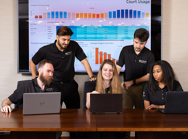 5 students are gathers around laptops working on a group project.