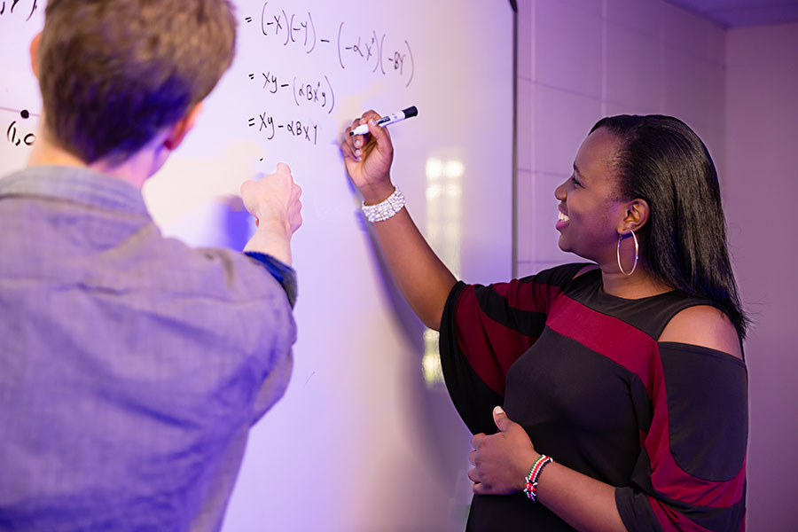 Math student and faculty working on research / A mathematics student writing an equation on a white board while a faculty member looks over.