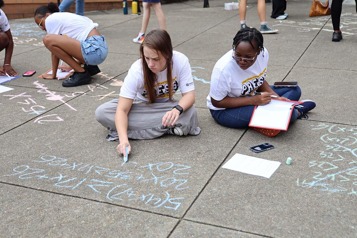 PRESS students from the college of science and mathematics sit on the pavement, writing math equations with chalk as part of an engaging group activity. / PRESS students from the college of science and mathematics sit on the pavement, writing math equations with chalk as part of an engaging group activity.