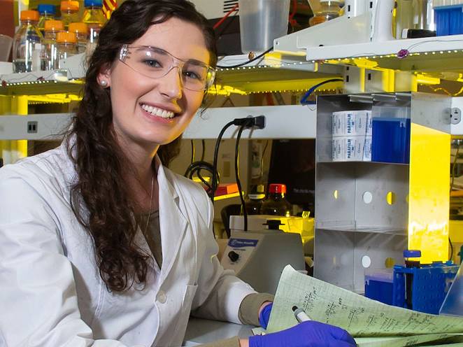 Women is wearing safety goggles and gloves sitting taking notes beside science equipment