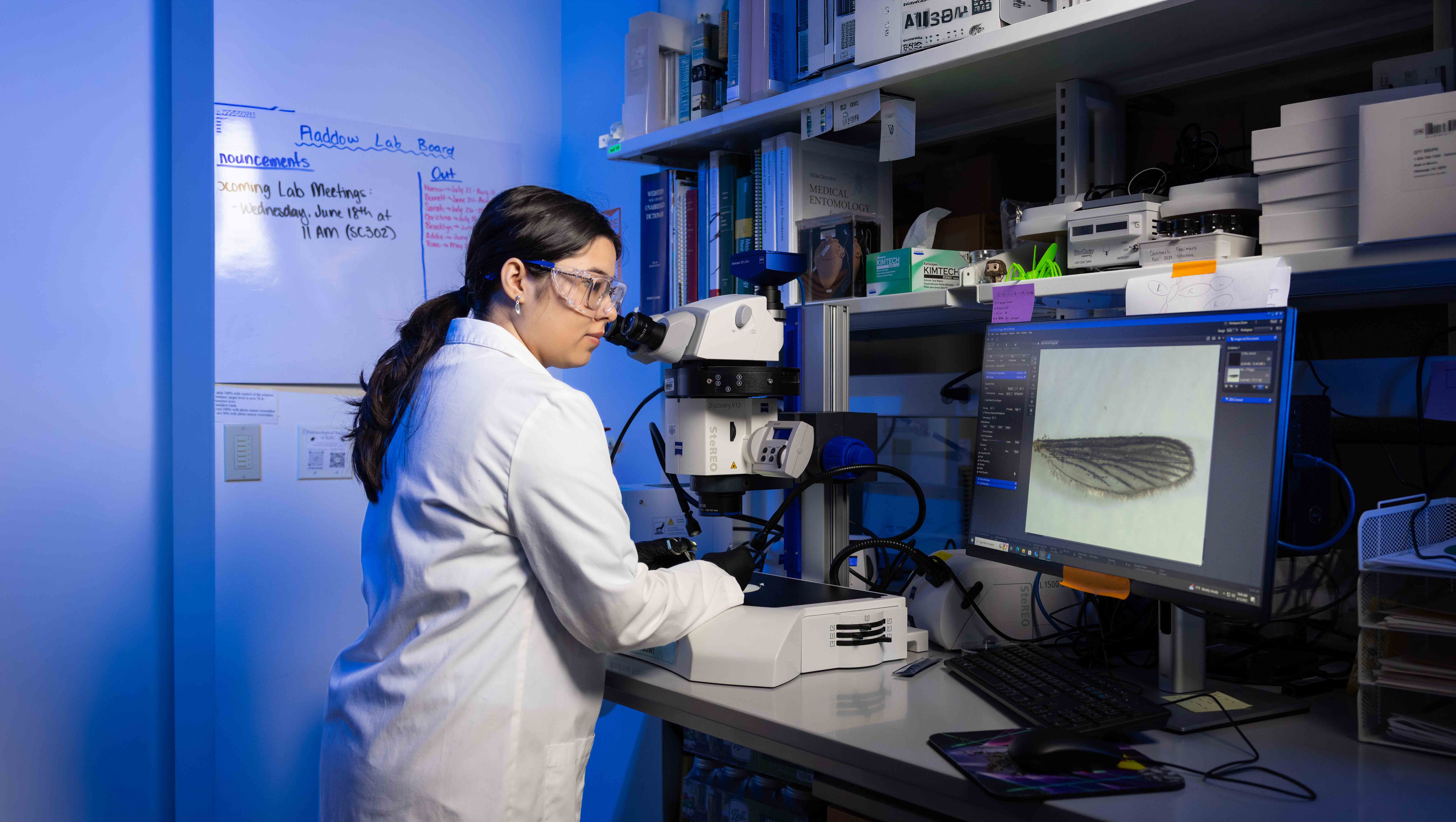 Laboratory researcher wearing protective eyewear and a lab coat operates a microscope in a science lab, surrounded by research materials, textbooks, and laboratory equipment.