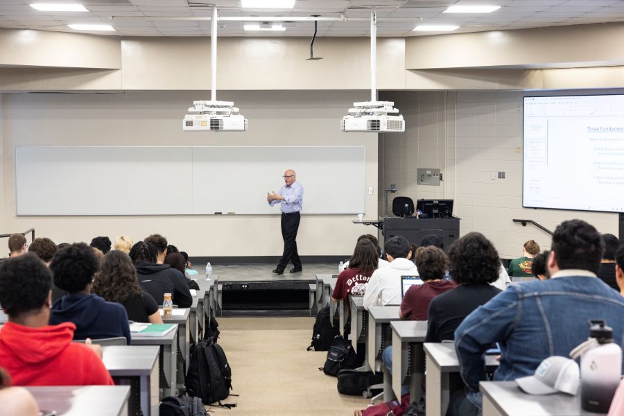 Classroom with a lecturer speaking to students, some using laptops, with a screen displaying text