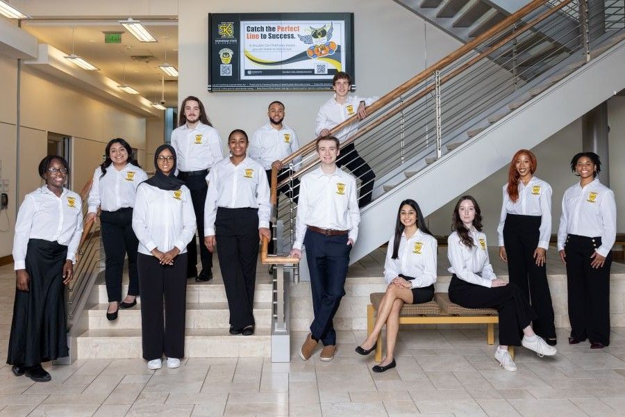 Group of people in white shirts and black pants posing indoors on stairs and a bench