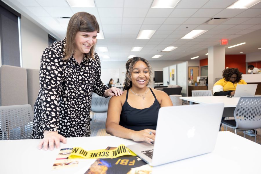 Two people in an office, one seated with a laptop and one standing, with others working in the background