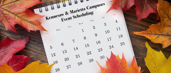 A monthly calendar page resting on a dark wooden surface. The calendar has black text and numbers. "Kennesaw & Marietta Campuses Event Scheduling" is printed in a black, stylized font across the top of the calendar page.