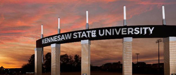 A large entrance sign for Kennesaw State University stands against a dramatic sunset sky. The sign features bold white lettering on a black curved arch supported by tall brick columns.