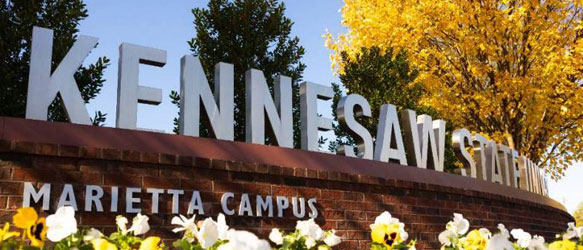 The entrance sign to Kennesaw State University’s Marietta Campus, featuring large silver letters mounted on a red brick wall. Colorful flowers bloom in the foreground, and trees with vibrant yellow autumn leaves frame the background.