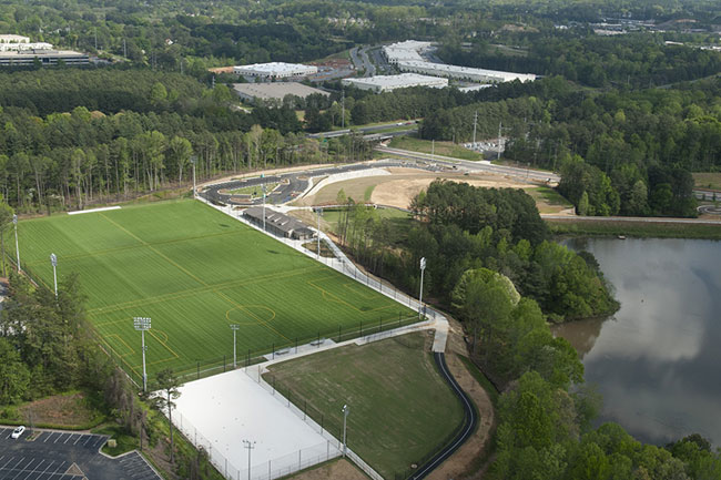 An aerial view of a large outdoor sports complex surrounded by trees and greenery. The complex includes multiple facilities such as a large green field and a smaller white rectangular area, with pathways, parking areas, and a nearby body of water.