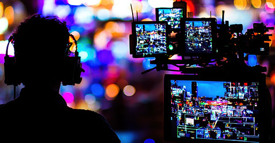 The photo is of a person wearing headphones, silhouetted against a colorful, blurred background of lights. In front of the person is a professional camera setup with multiple screens.