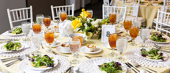 A KSU Elegant round banquet table set for a formal event with white chairs, iced tea, water glasses, fresh salads, and floral centerpieces in a bright and cheerful setting.