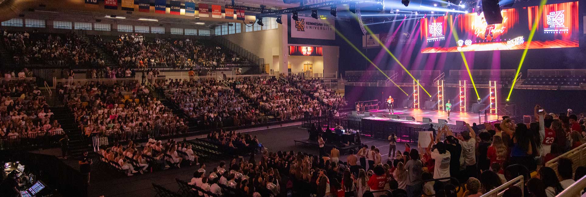 KSU's indoor stadium filled with a seated audience watching a live performance on stage, with bright spotlights and Kennesaw State University banners displayed overhead.