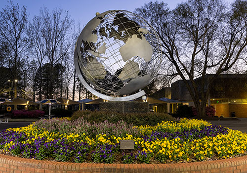 A large metal globe sculpture on the Kennesaw State University Marietta campus, set in the middle of a circular brick flower bed filled with vibrant yellow and purple flowers. The globe is surrounded by a well-maintained walkway, with trees and campus buildings visible in the background during dusk, adding a peaceful ambiance to the scene.