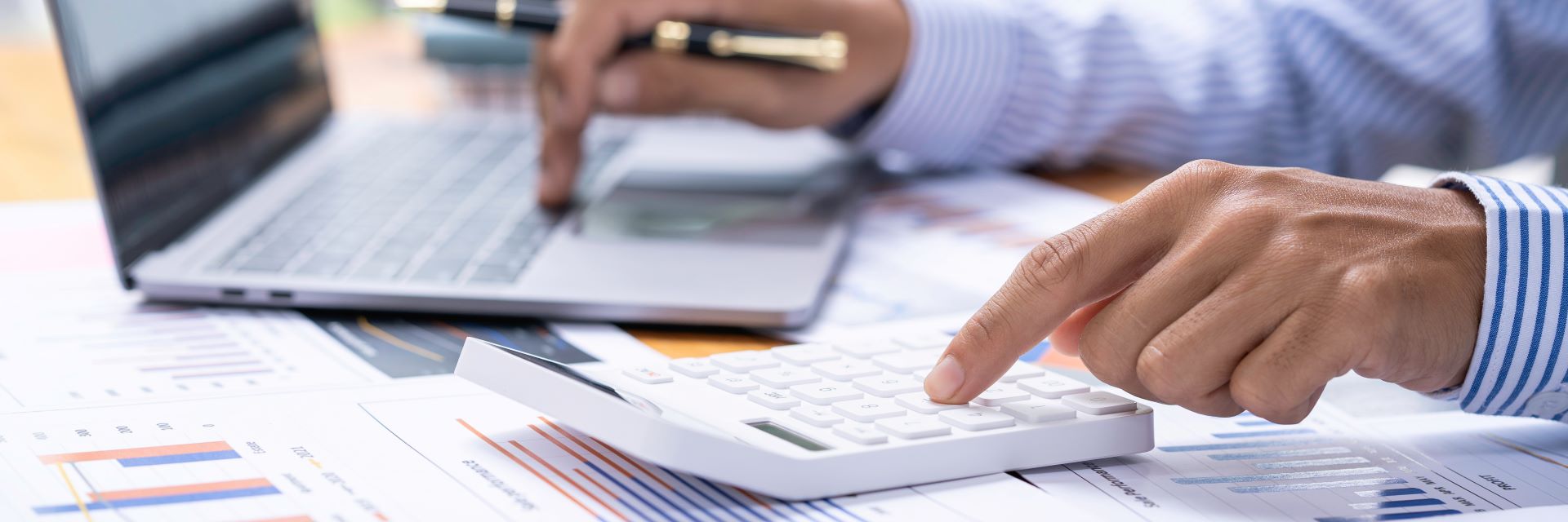 Accountant using a calculator and laptop, surrounded by forms with charts and graphs