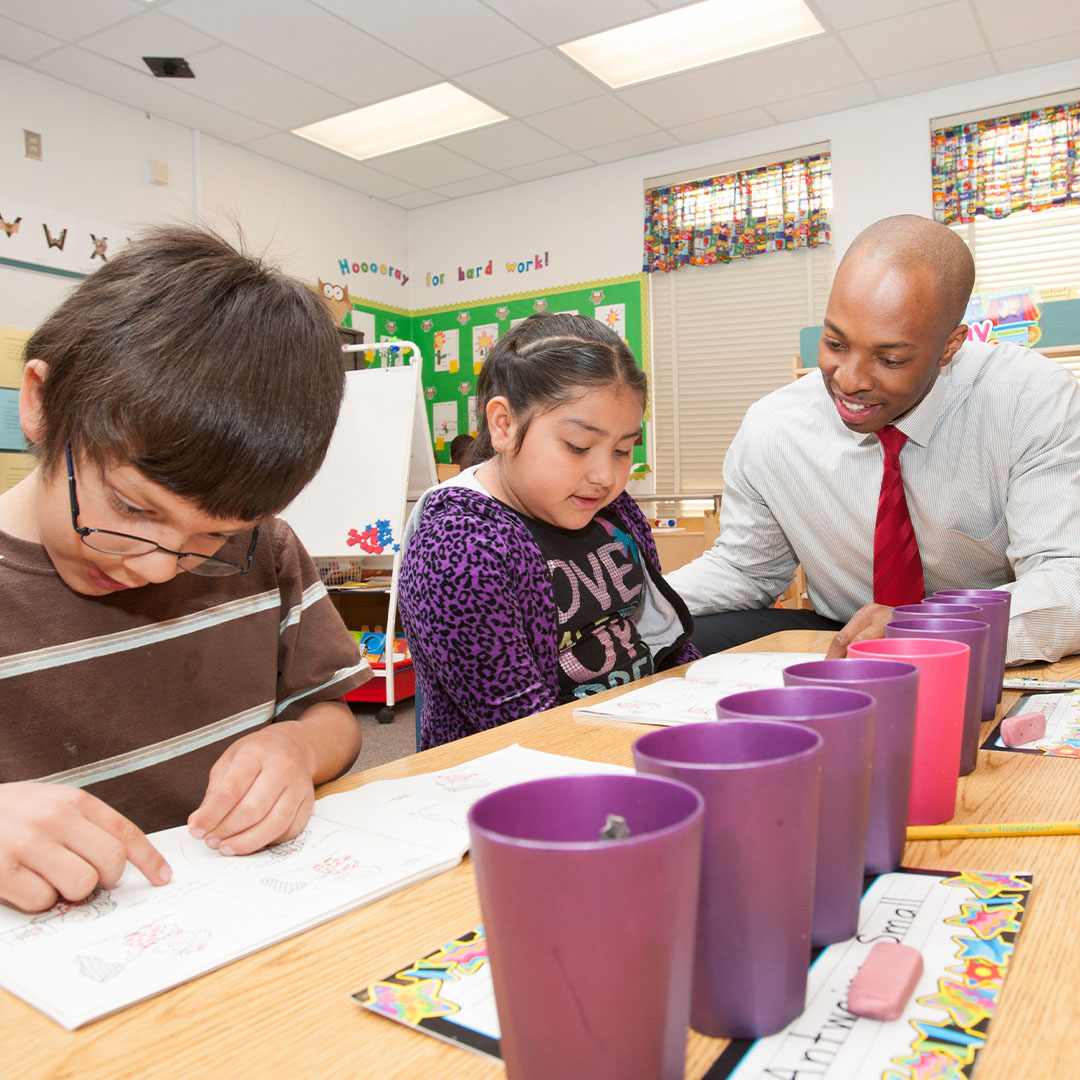 A teacher sits at a table with two young students who are working in activity books. Several purple plastic cups are lined up on the table, and the classroom is decorated with colorful posters and materials.