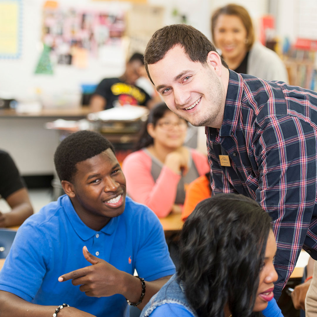 A classroom with older students seated at desks talking with an instructor who is leaning in to engage with them. Other students work or talk in the background.