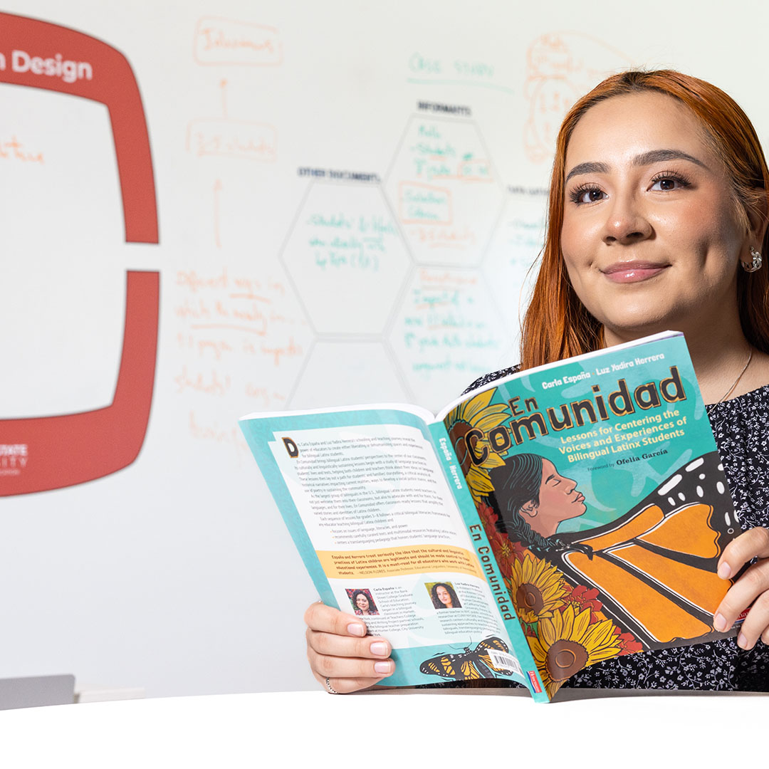 A person holds open a colorful book titled “En Comunidad” while sitting in front of a whiteboard covered in diagrams and notes. The book cover features illustrations of sunflowers and butterflies.