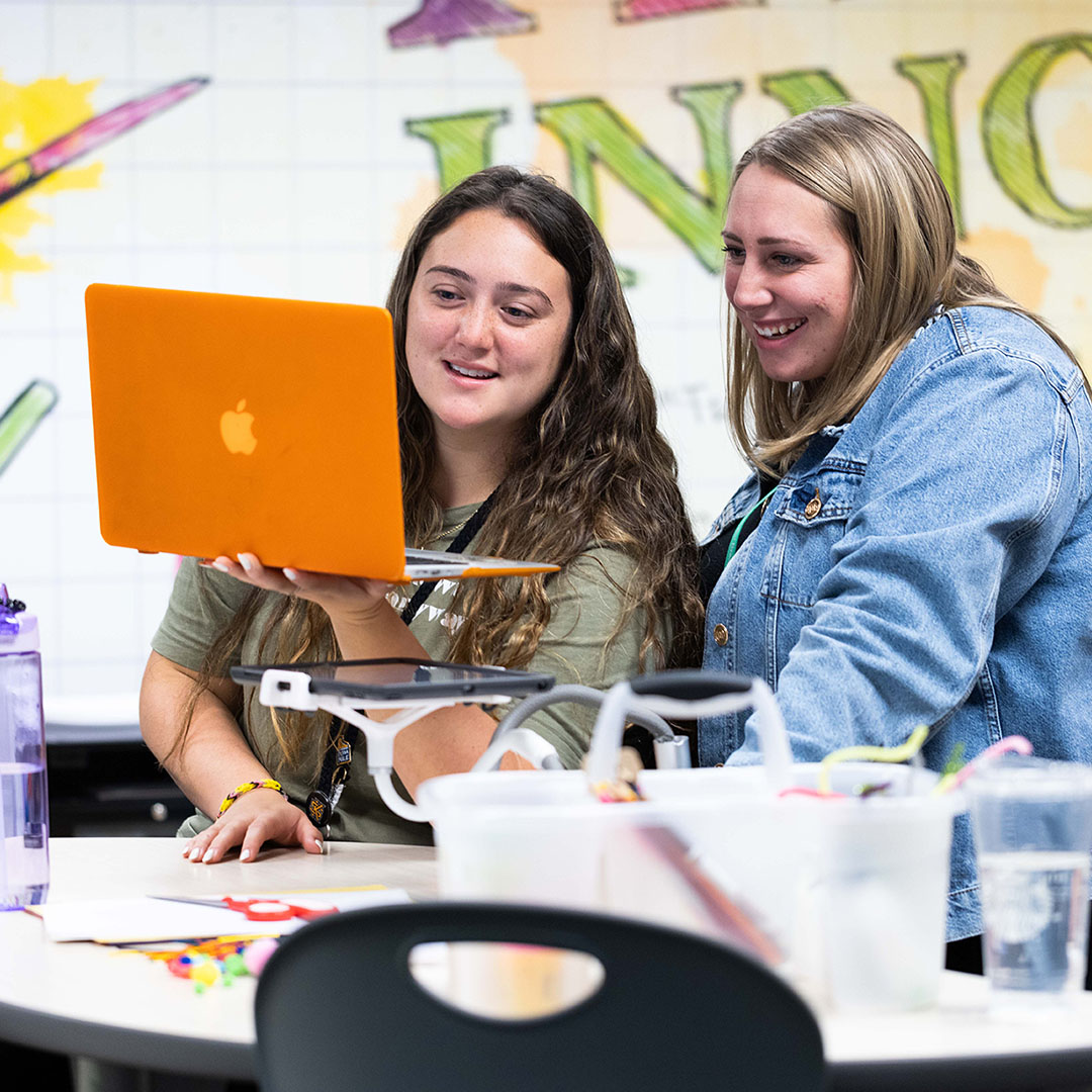 Two individuals reviewing information on an orange laptop at a classroom table, surrounded by instructional materials and supplies.