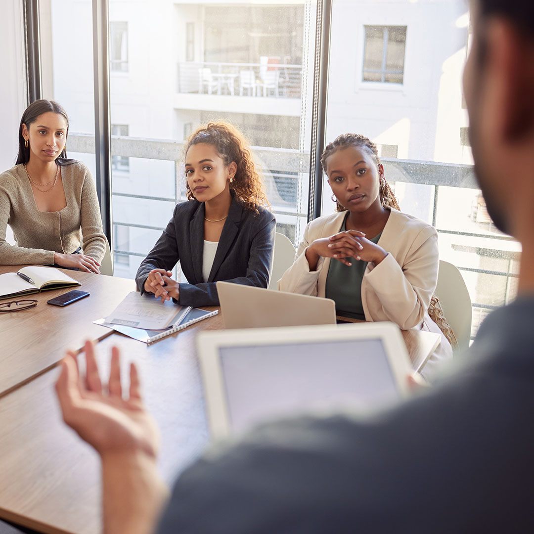  professional leads a discussion during a meeting, addressing colleagues seated at a conference table with laptops and notes in a modern office setting.