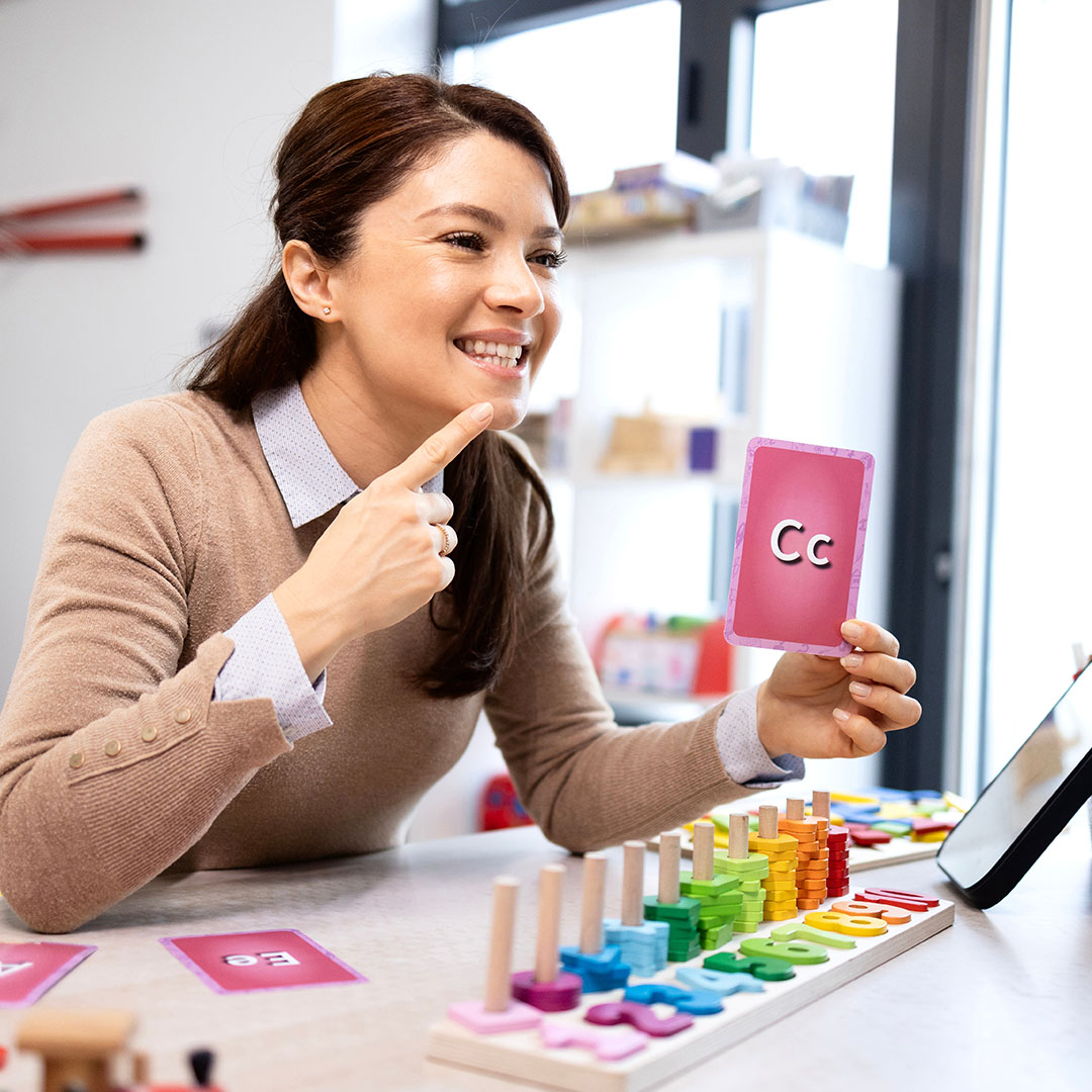 A professional demonstrates an instructional activity using a phonics flashcard labeled “Cc,” with educational materials and learning tools arranged on a table.