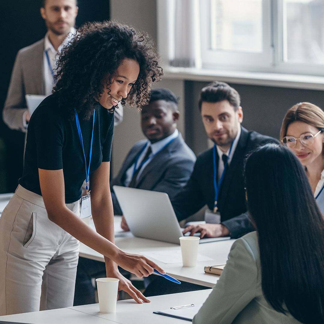 A professional collaborates with colleagues during a training session, pointing to materials on a table while others follow along using laptops in a classroom-style environment.
