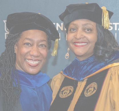 Two women dressed in graduation regalia pose happily for a photo, showcasing their achievements.