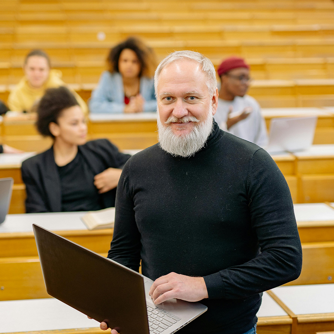 College professor smiling while holding laptop and students behind him
