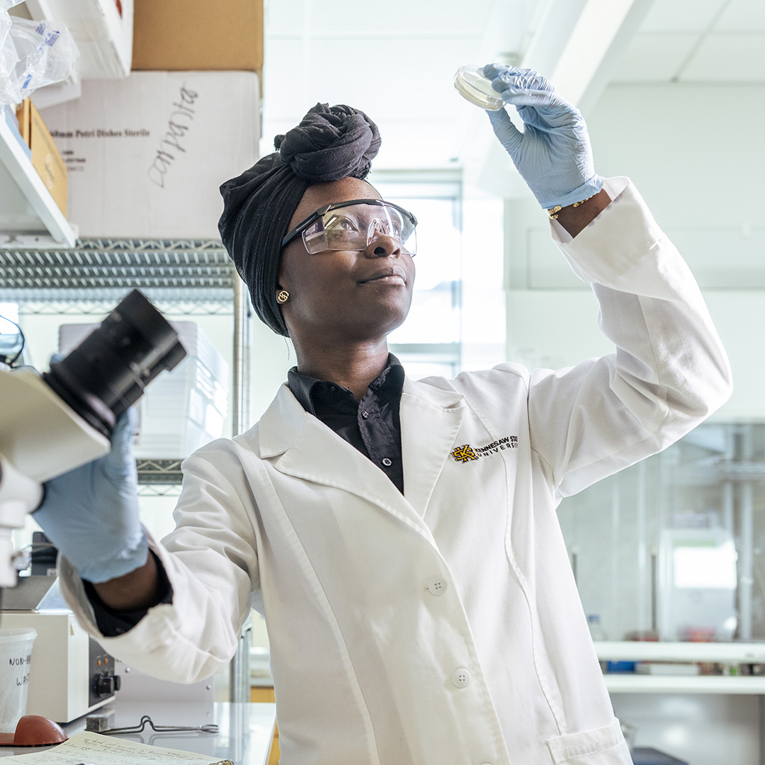 A woman in a lab coat examines a petri dish in her hand, standing in a laboratory setting.