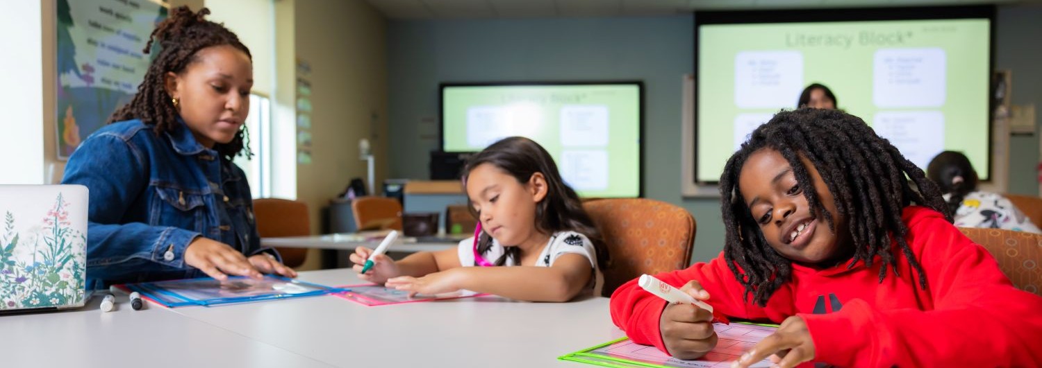 Two children write on laminated sheets with dry-erase markers as a teacher assists them in a classroom