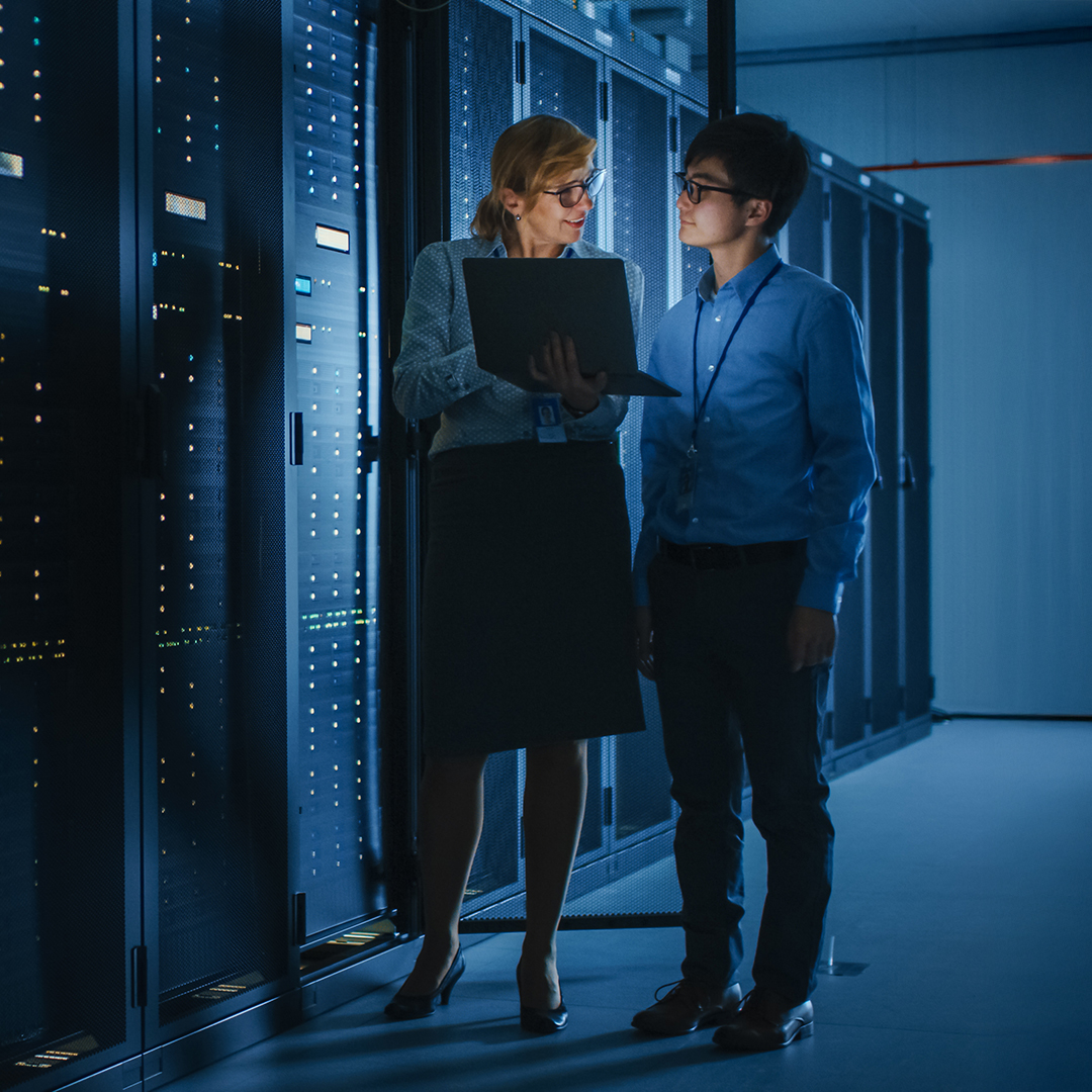 Two people standing in front of tall servers in a data center, discussing technology and data management.