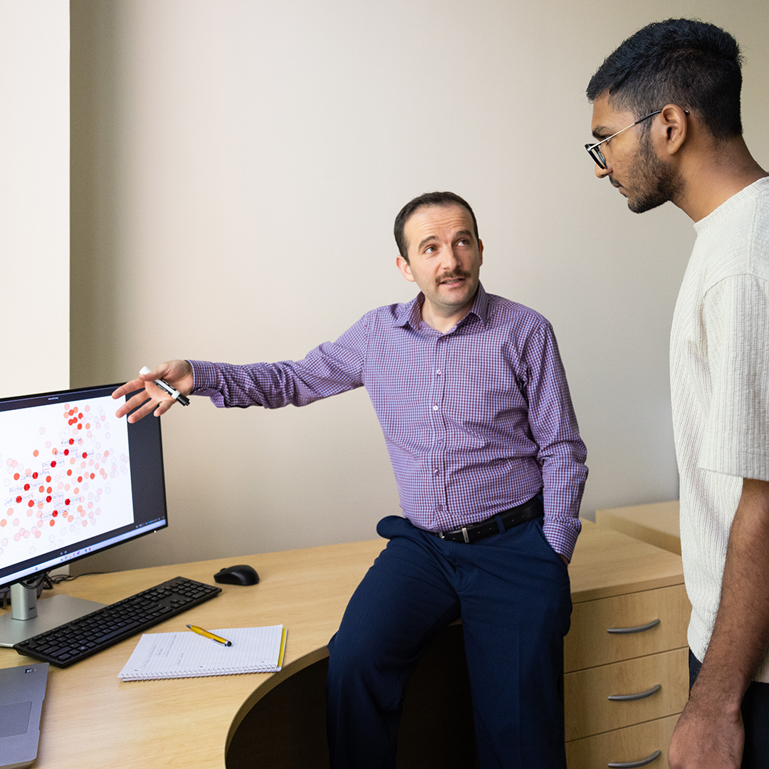 A man points at a computer screen while another man stands beside him, both engaged in discussion.