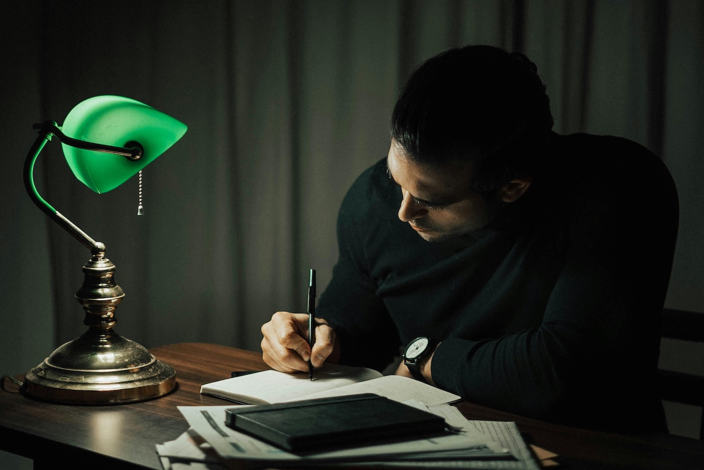 A man is focused on writing at a desk illuminated by a desk lamp.
