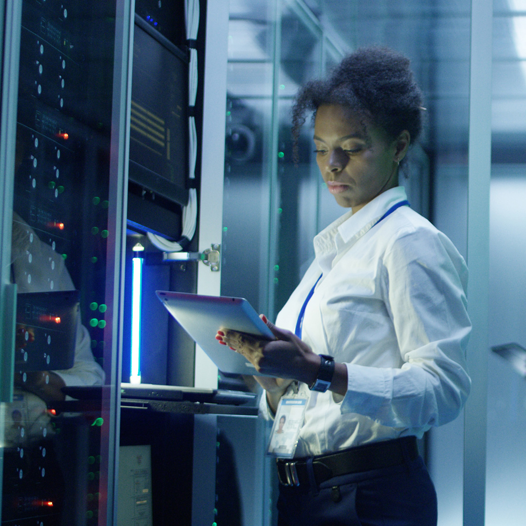 A woman stands in a server room, holding a tablet while monitoring the servers.