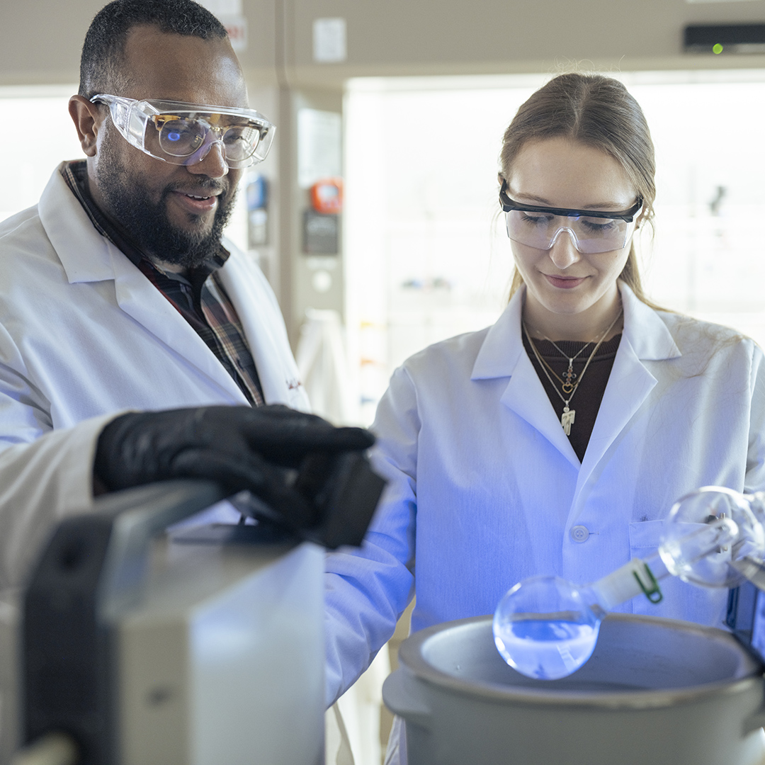 Two researchers in lab coats conduct a chemistry experiment using specialized laboratory equipment, focusing on a glass flask containing a blue liquid in a modern scientific lab setting.