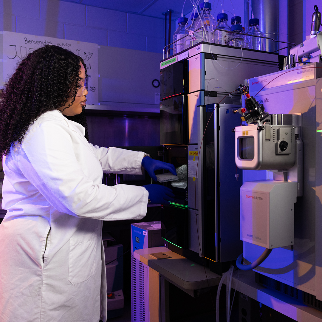 A laboratory technician in a white coat operates advanced analytical equipment in a high‑tech forensic science lab, working with illuminated instruments and specialized testing systems.