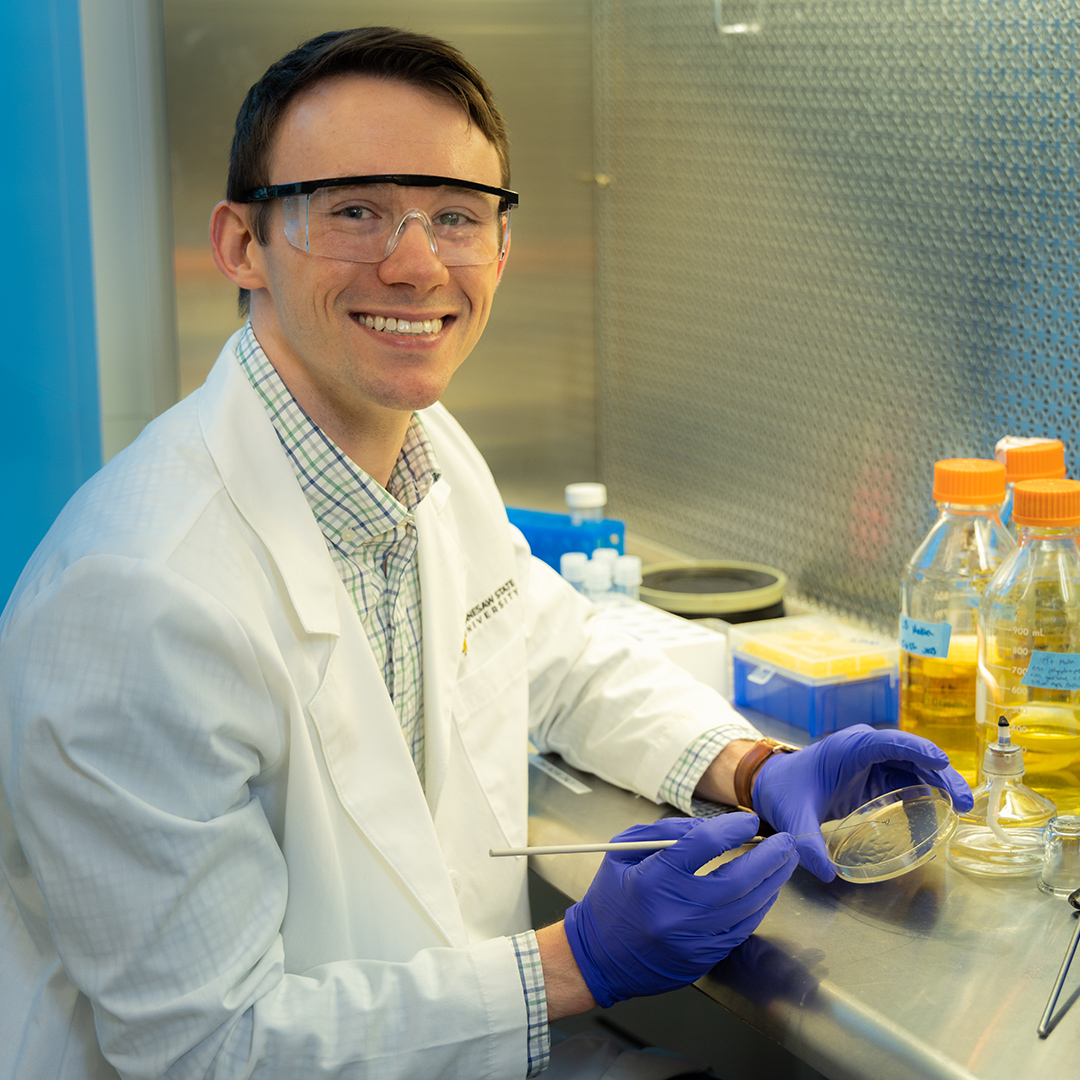 A laboratory researcher in a white coat works with a petri dish inside a sterile lab environment, using purple nitrile gloves and specialized equipment to conduct scientific analysis.