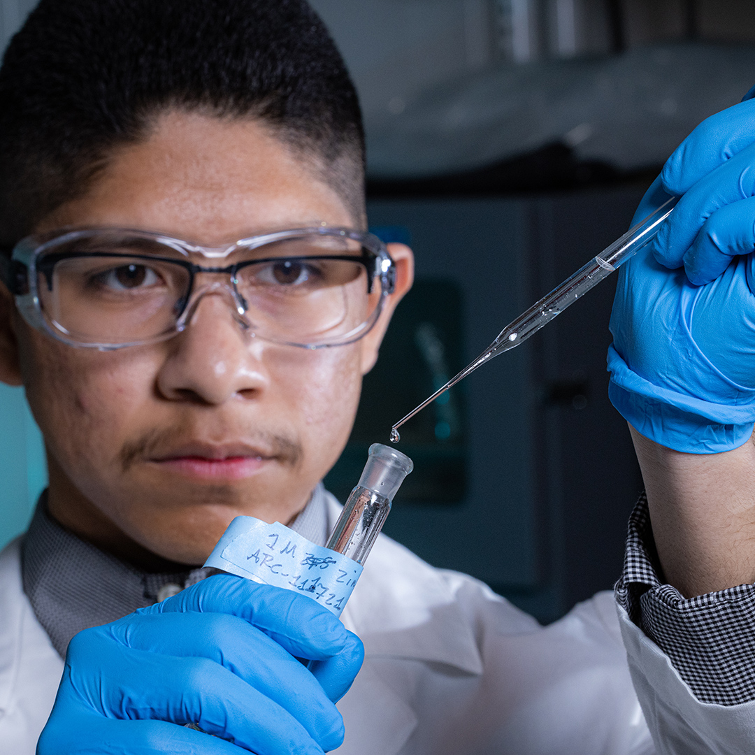 A scientist wearing blue gloves carefully transfers liquid with a pipette into a labeled container during a laboratory experiment.