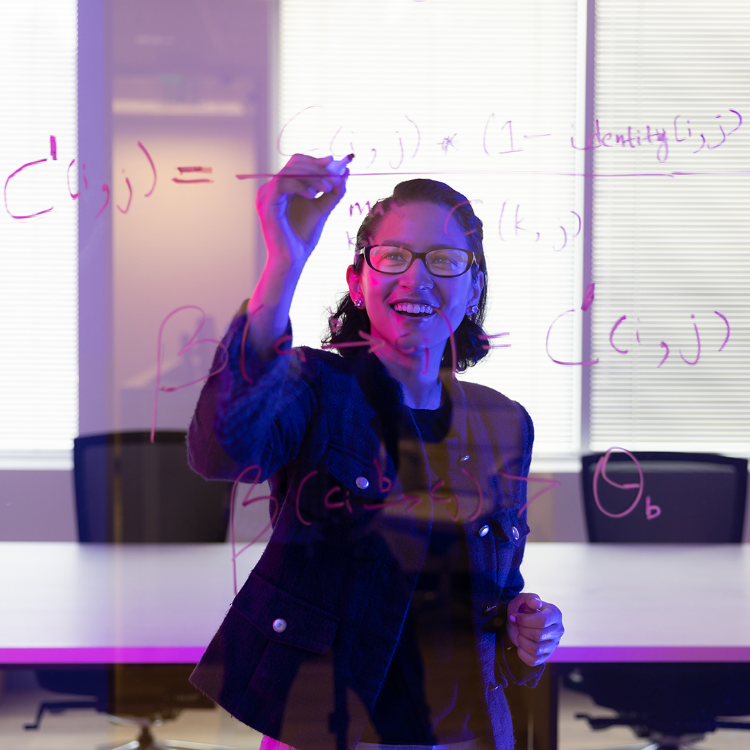 A woman with glasses is actively writing mathematical equations on a see-through clear board.
