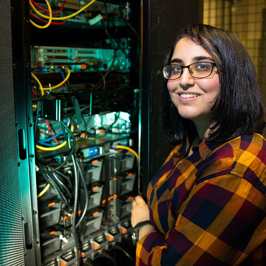 A woman in a plaid shirt and glasses poses in front of a server, highlighting her role in a tech environment.
