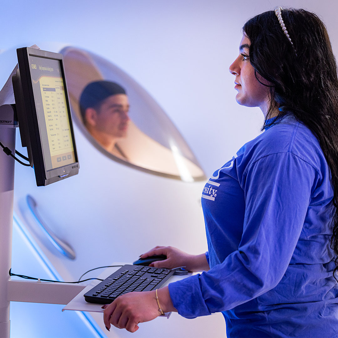 Student operating a computer to record body composition data during an assessment using advanced measurement equipment in a clinical exercise science laboratory.